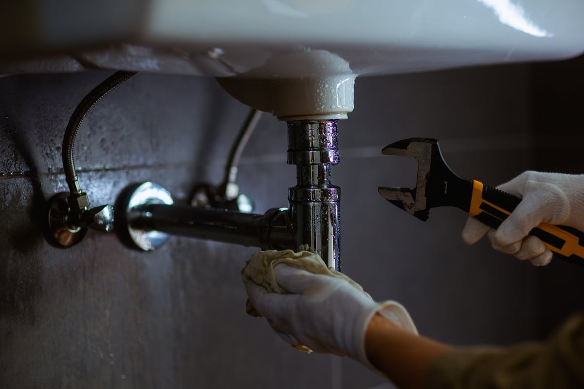 Someone in white gloves works on plumbing under a white sink using an adjustable wrench and a rag, possibly DIY home repair in a bathroom.